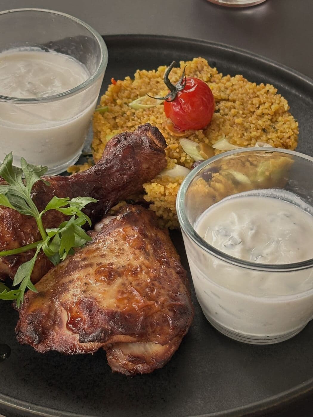 Hähnchenkeulen auf Couscous mit Kirschtomate, dazu zwei Gläser mit hellem Dip