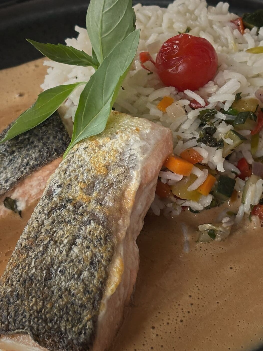 Nahaufnahme: gebratenes Fischfilet mit Haut auf Gemüsereis, heller Sauce, Kirschtomate und Basilikum