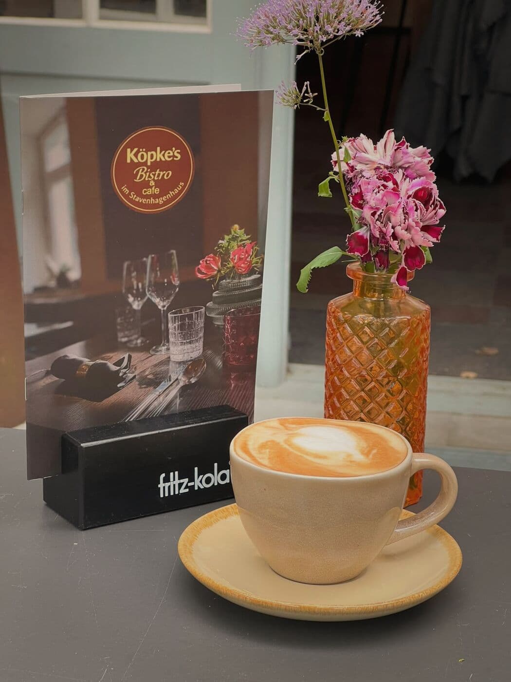 Cappuccino mit Milchschaum-Herz auf einer Untertasse, daneben eine Vase mit Blumen und die Köpke's-Karte